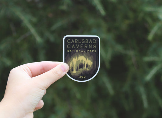 A hand holds the Carlsbad Caverns National Park sticker, its lit cave formations and 1930 date set against green pine.
