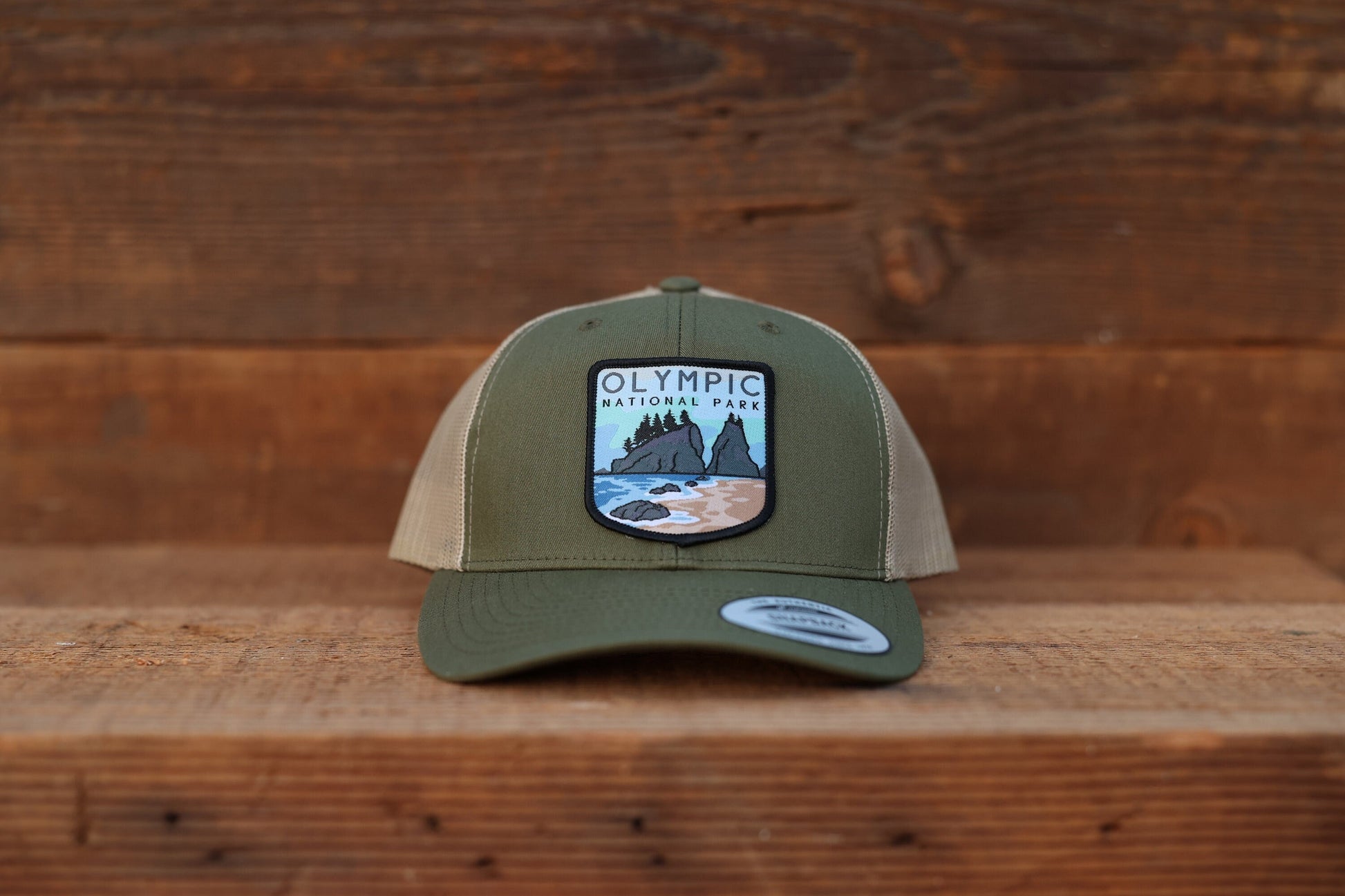 Squared to the camera on plank wood, the olive Olympic National Park trucker hat shows its tan side mesh.