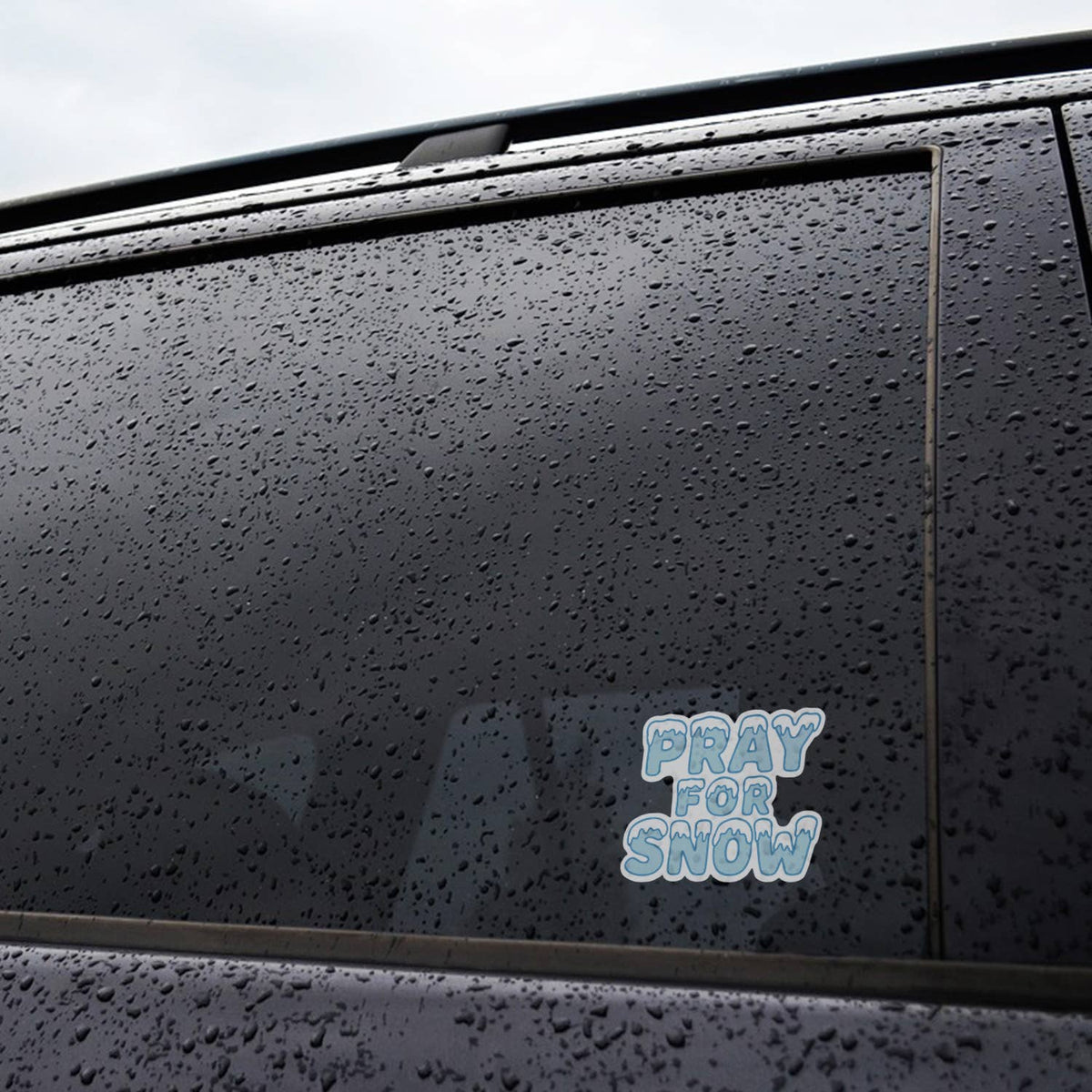 Rain beads across a black car door where the Pray For Snow sticker sits near the bottom of the window.