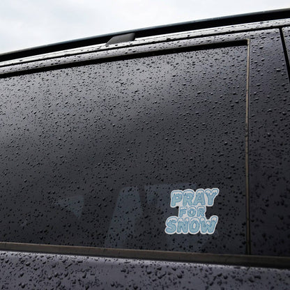 Rain beads across a black car door where the Pray For Snow sticker sits near the bottom of the window.