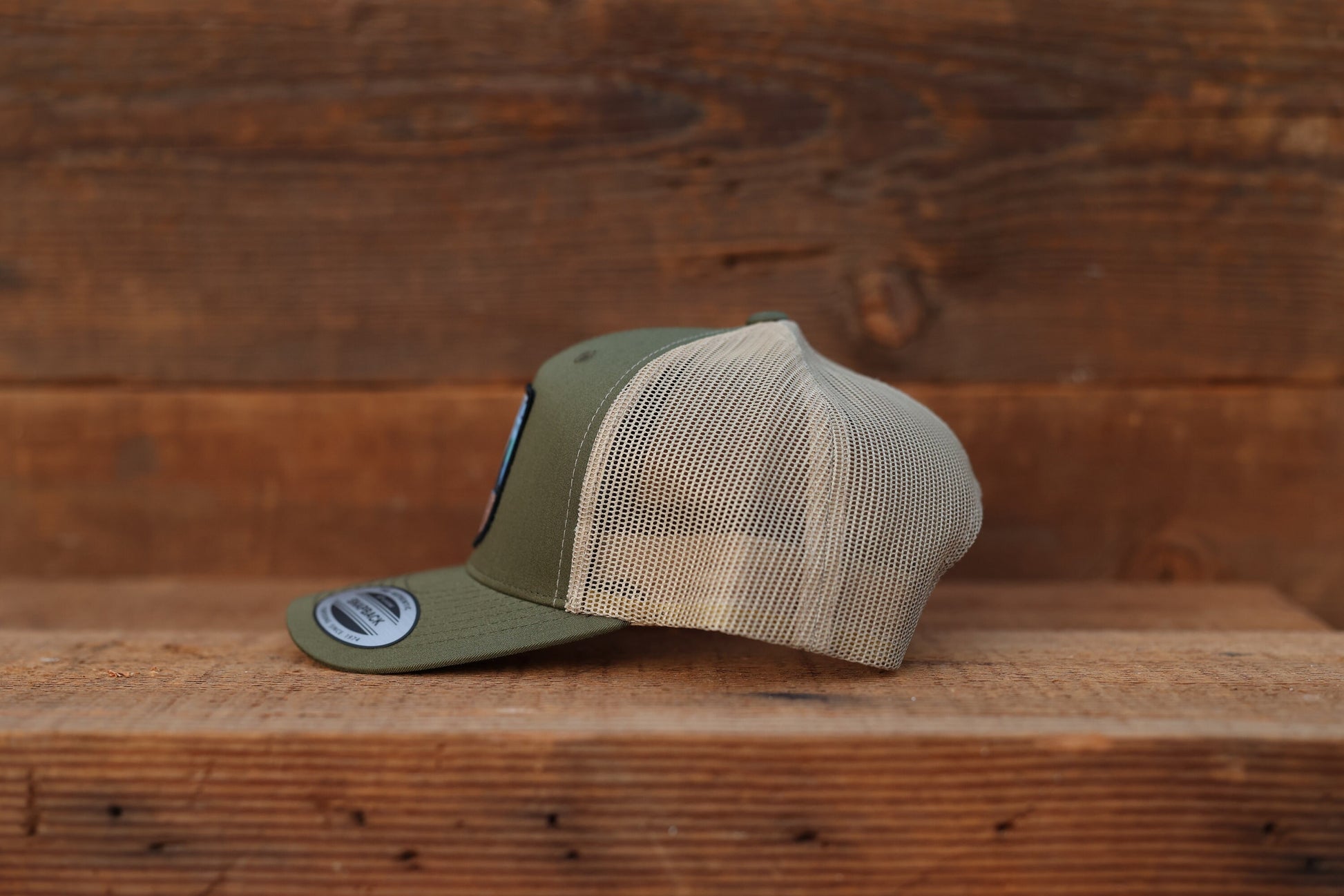 Seen from the left on a wood shelf, the olive Olympic National Park trucker hat shows tan mesh panels.
