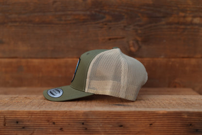 Seen from the left on a wood shelf, the olive Olympic National Park trucker hat shows tan mesh panels.