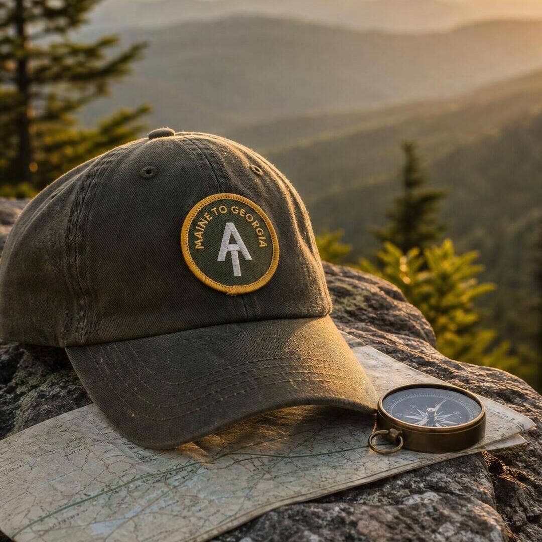 Stitched to a faded green cap on a rock ledge, the AT Maine to Georgia patch sits by a compass and map.