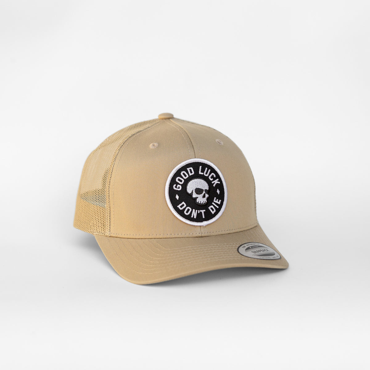 Tan canvas covers the entire Good Luck Don't Die trucker hat, the skull patch standing out in black and white.
