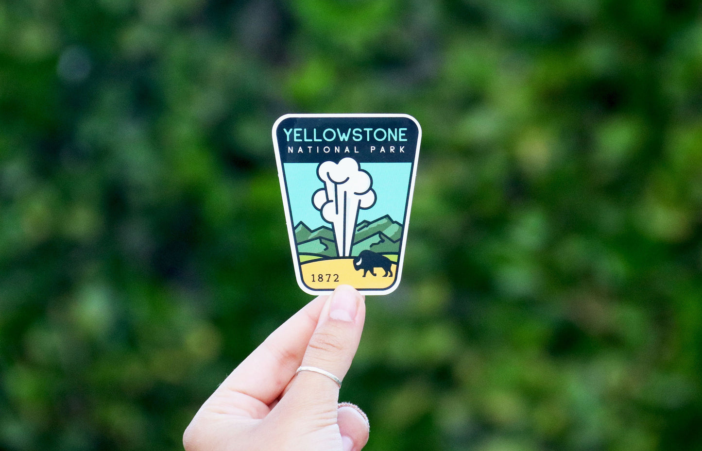 Held between two fingers, the Yellowstone National Park geyser sticker stands against soft green leaves.