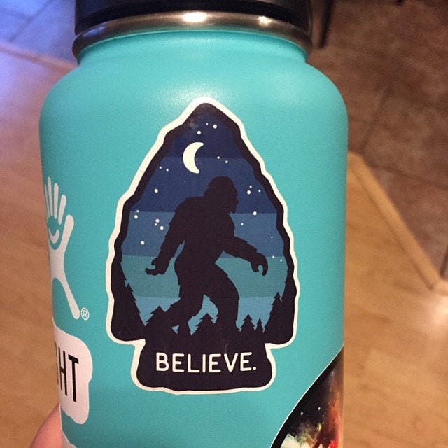 A teal water bottle wears the Bigfoot Believe sticker beside other decals on a tile kitchen floor.