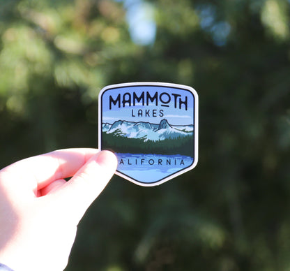 Shaded by trees, the Mammoth Lakes sticker sits pinched between finger and thumb with California lettering showing.