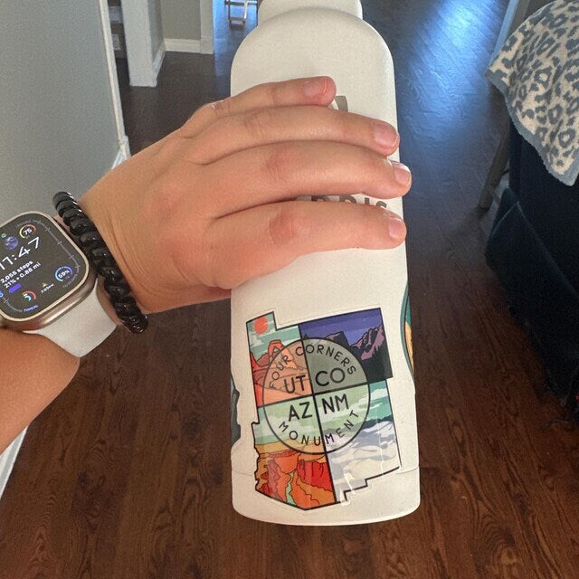 A hand grips a white water bottle indoors where the Four Corners Monument sticker sits among other stickers.