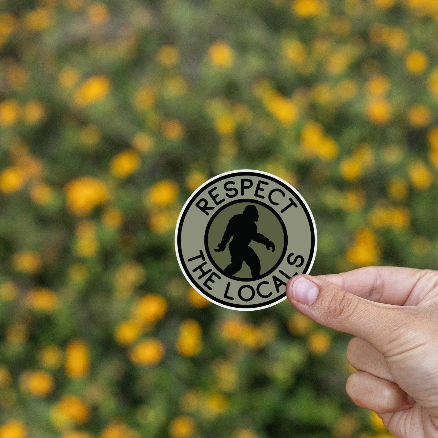 Yellow wildflowers blur behind fingers pinching the Bigfoot Respect The Locals sticker.