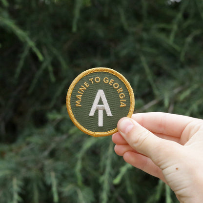 A hand holds the AT Maine to Georgia patch in front of dense evergreen branches.