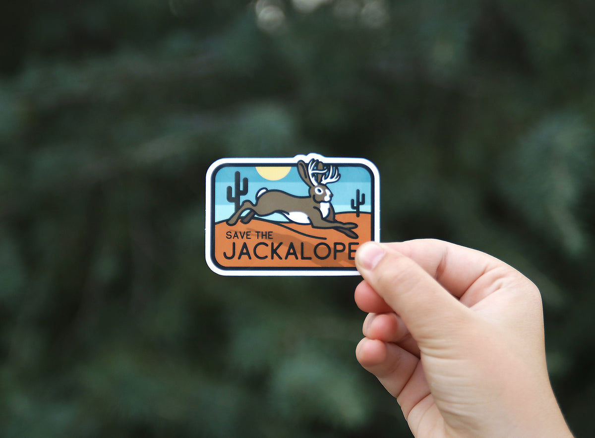 Backed by a blurred evergreen, the Save The Jackalope sticker sits small between finger and thumb.