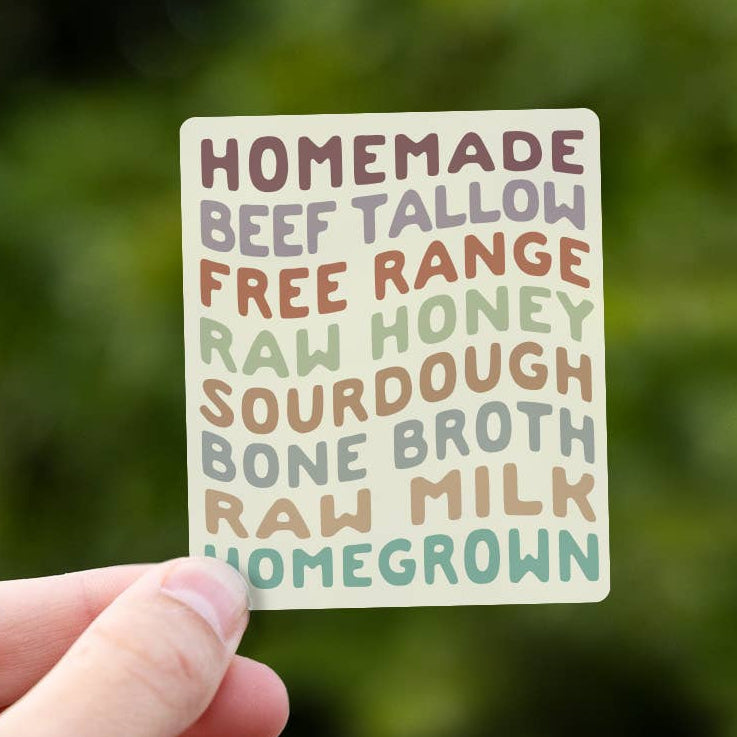 A hand holds the Homemade Essentials sticker against blurred green leaves, its stacked food words in muted colors.