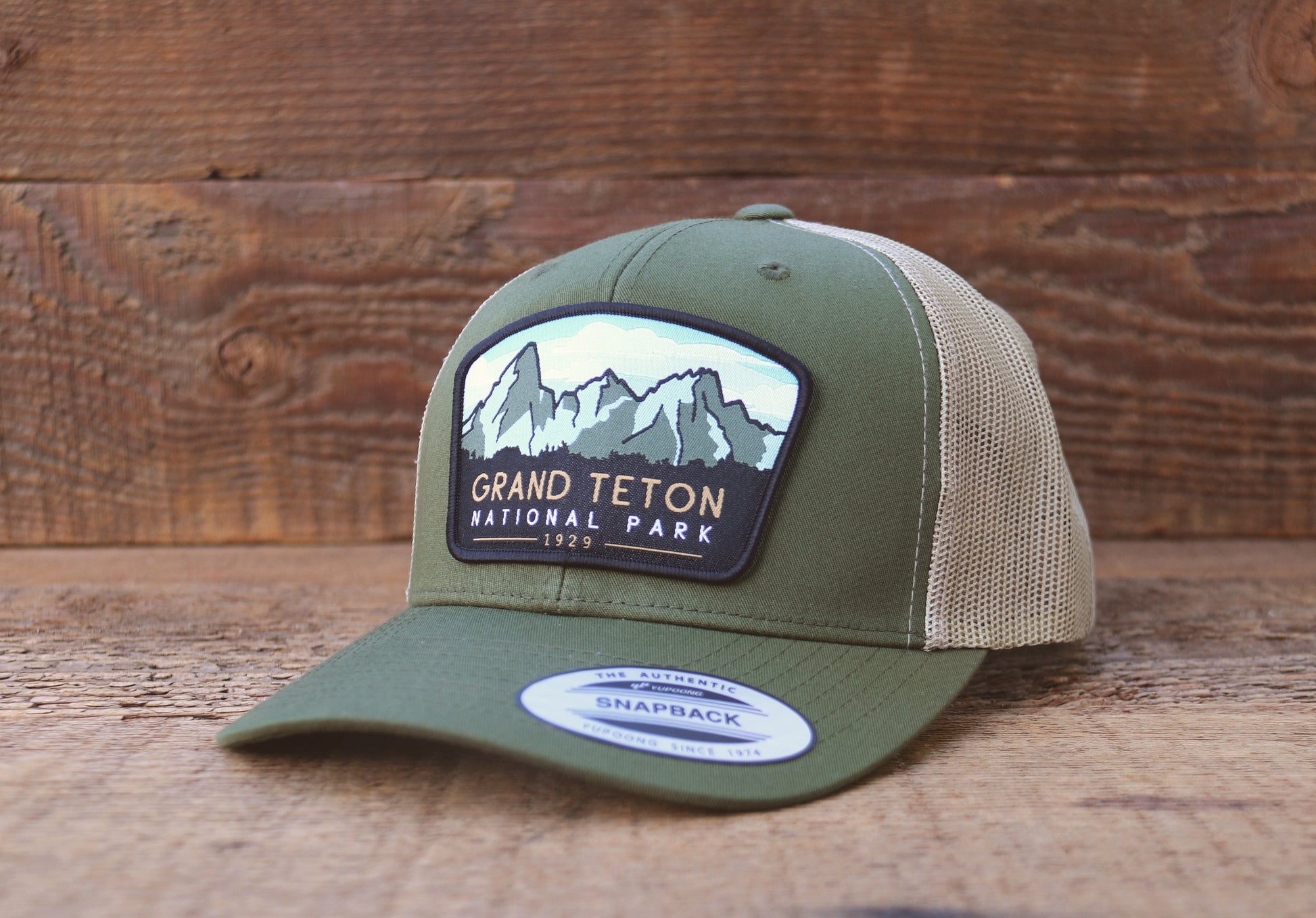 Set on weathered wood planks, the olive Grand Teton National Park trucker hat still wears its snapback sticker.