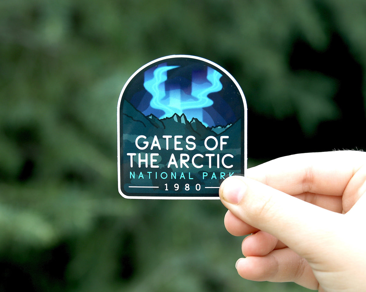 A hand holds the Gates of the Arctic National Park sticker, its aurora and peaks bright against blurred green boughs.