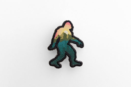 On a plain white surface, the Bigfoot Adventure Patch shows a sunset and pine forest inside its black outline.