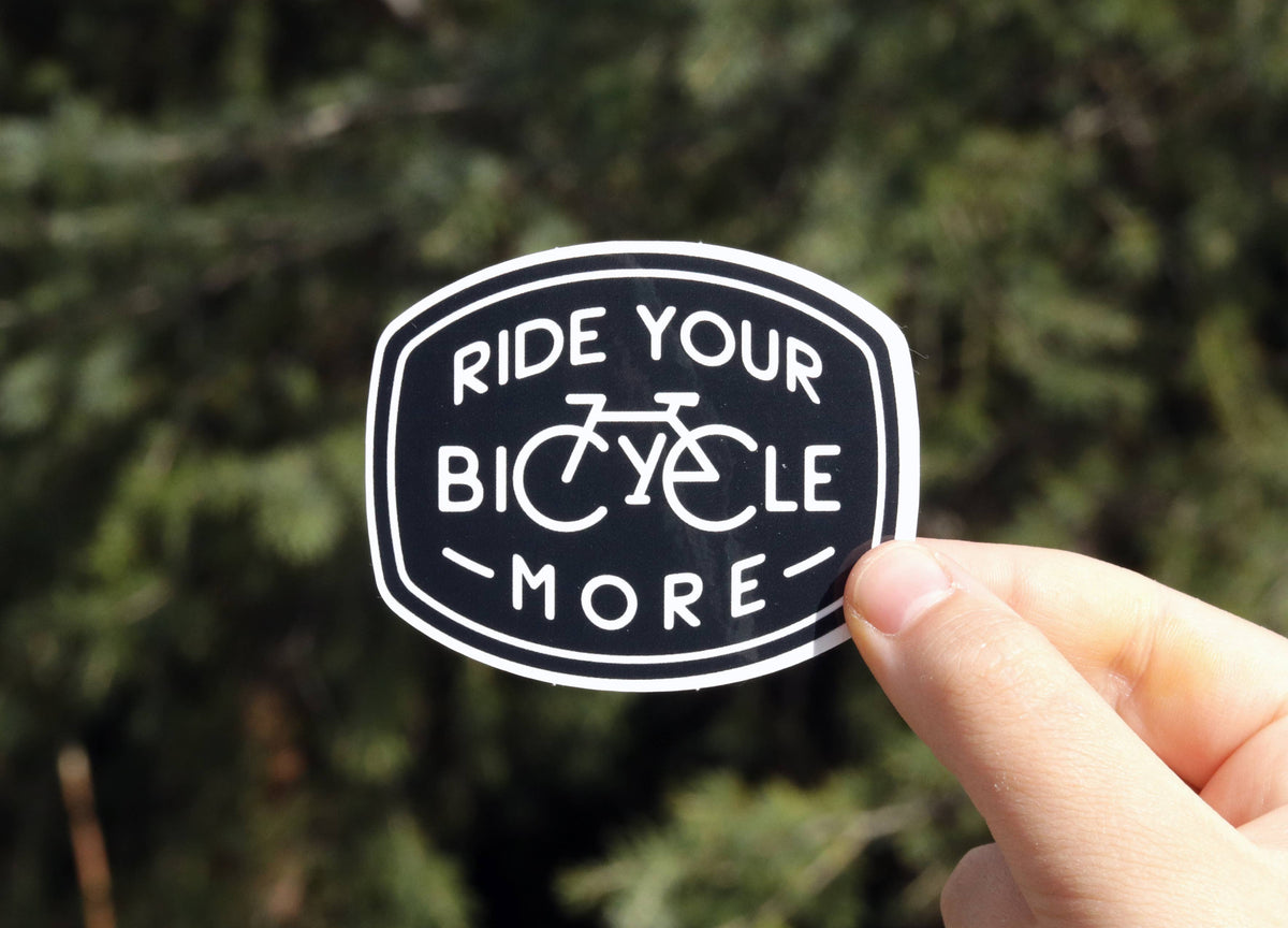 Held up against a blurred evergreen, the black Ride Your Bicycle More sticker shows its white badge lettering.