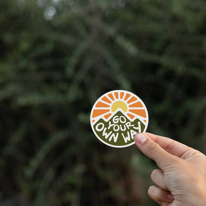 Pine branches sit out of focus behind the Go Your Own Way sticker held in one hand.