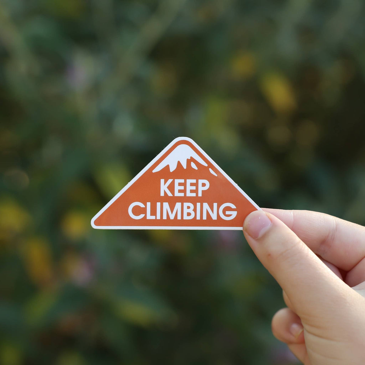 A hand holds the orange triangular Keep Climbing sticker with its snowy peak against blurred green brush.