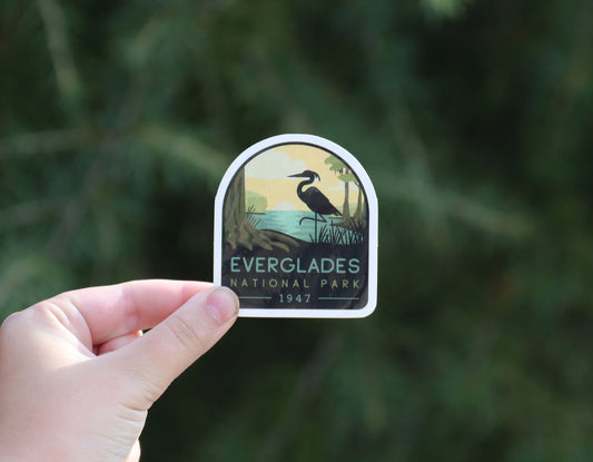 Held further back against blurred evergreen branches, the Everglades National Park sticker shows its 1947 date.