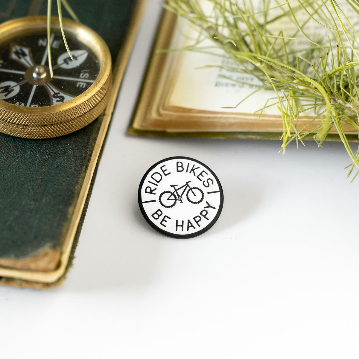 A compass and old book flank the Ride Bikes Be Happy pin resting beside green sprigs on a dark cover.