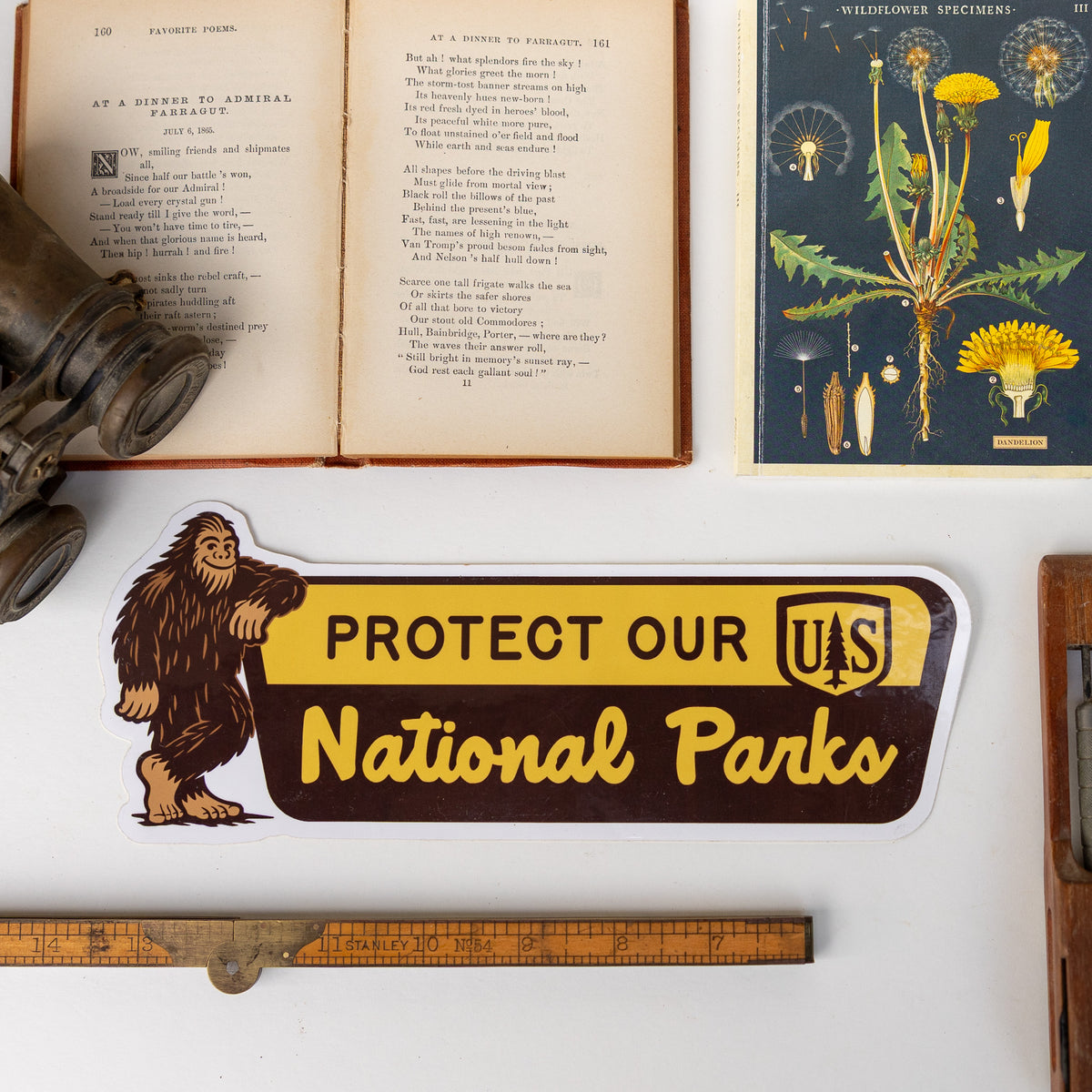 The Protect Our National Parks bumper sticker with Bigfoot rests near an open poetry book and wildflower print.
