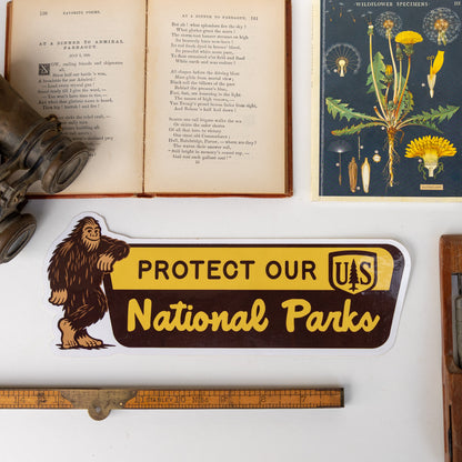 The Protect Our National Parks bumper sticker with Bigfoot rests near an open poetry book and wildflower print.