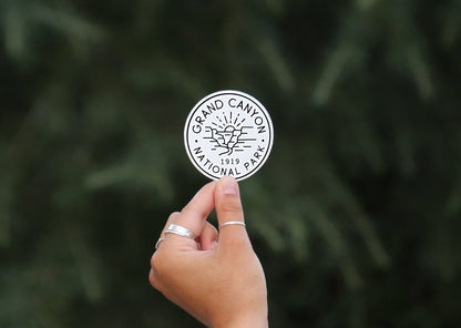 A ringed hand holds the black and white Grand Canyon National Park sticker against blurred evergreen branches.