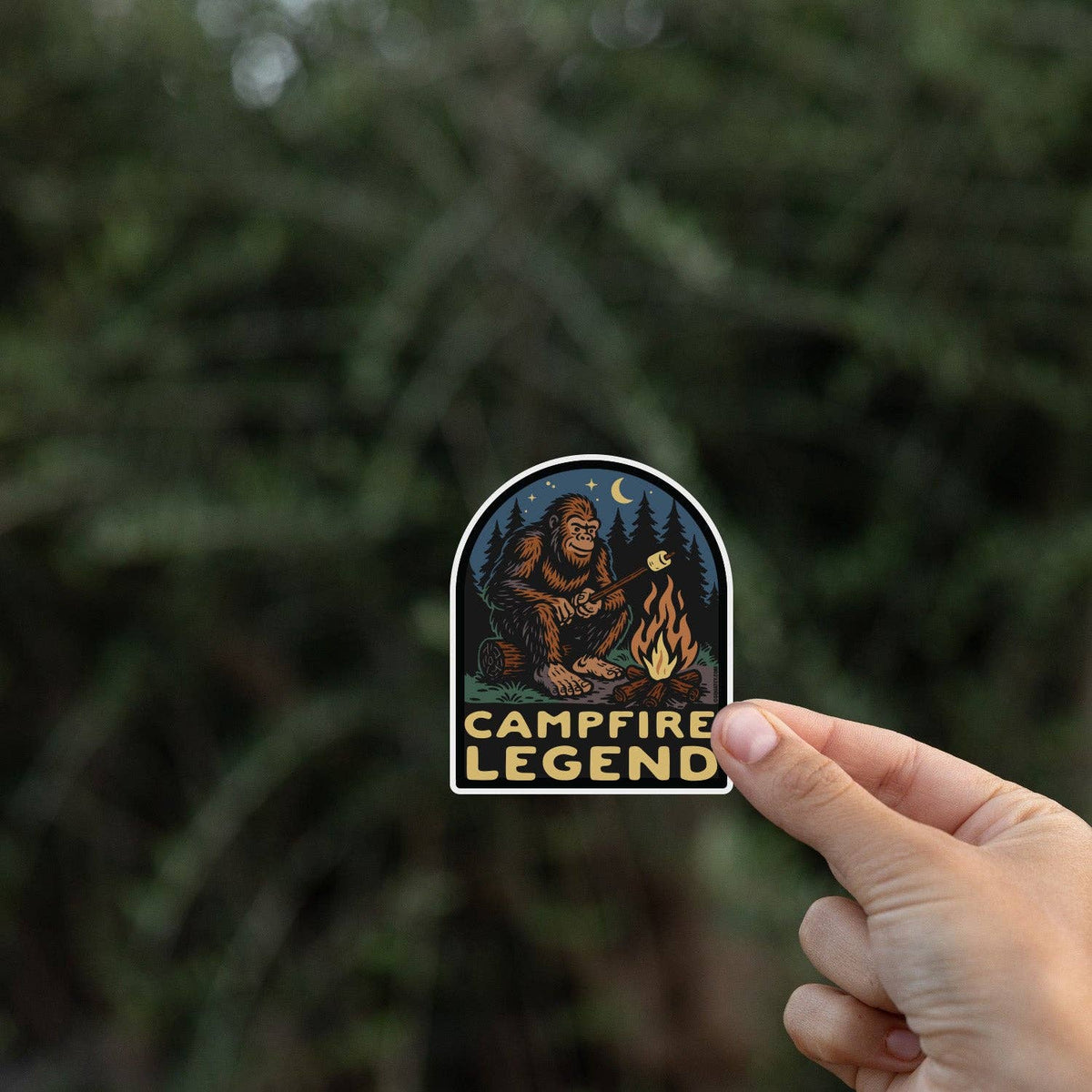 Fingers hold the Campfire Legend sticker in front of a blurred wall of dark evergreen branches.