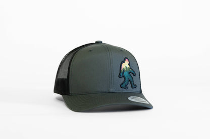 Charcoal Bigfoot Adventure trucker hat turned slightly right, its sunset colored Sasquatch patch catching the light.