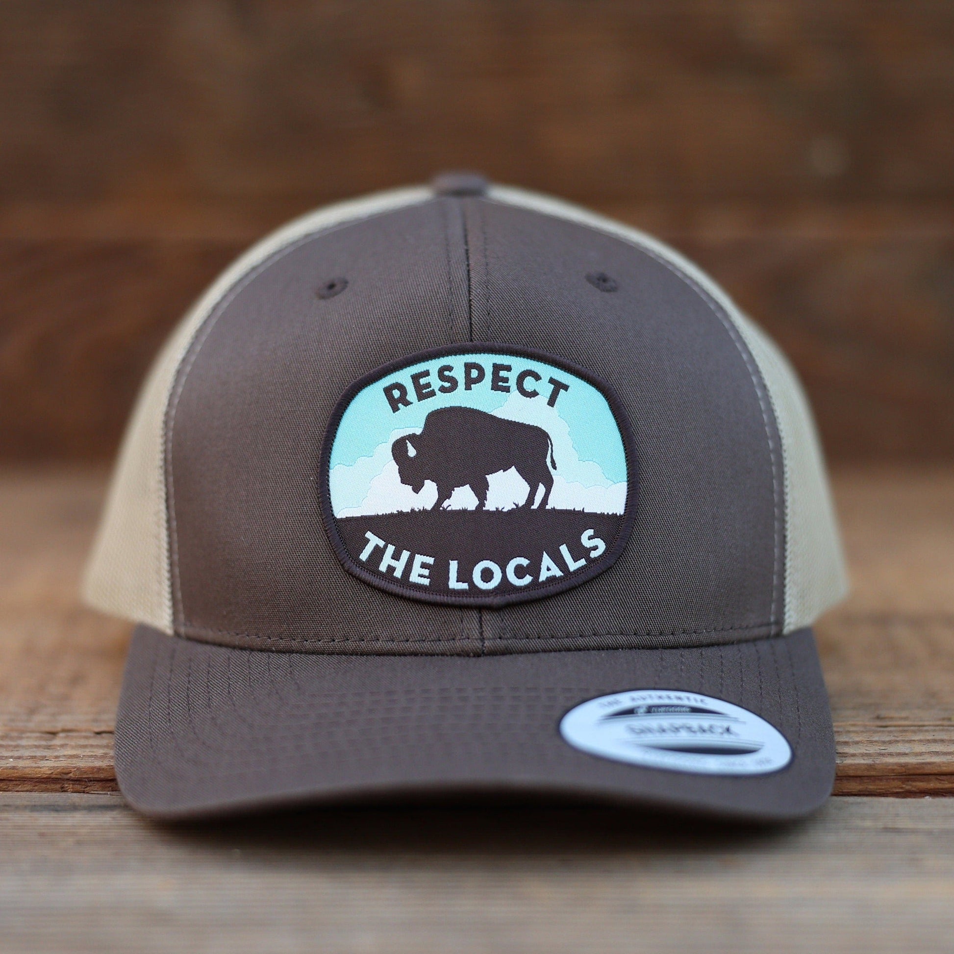 Straight on against weathered wood, the brown Respect The Locals Bison Trucker Hat centers its bison patch.