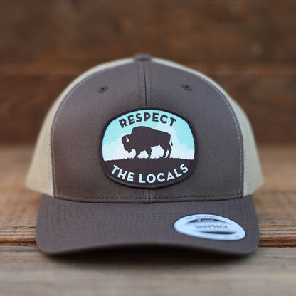 Straight on against weathered wood, the brown Respect The Locals Bison Trucker Hat centers its bison patch.