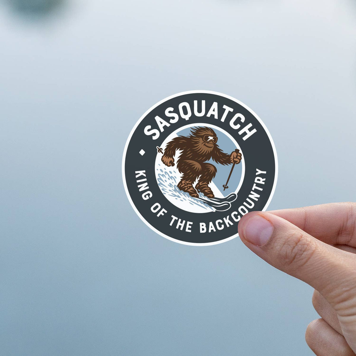 Fingers hold the Sasquatch King of the Backcountry sticker against a soft grey blue background.