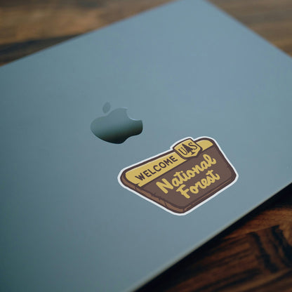 On the lid of a blue laptop beside the apple logo, the National Forest Sign sticker sits at an angle.