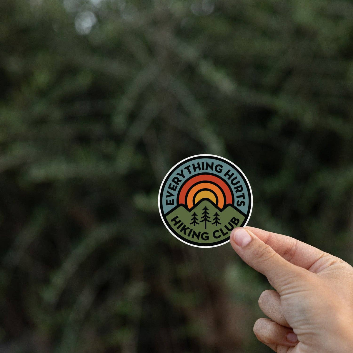 A hand pinches the Everything Hurts Hiking Club sticker in front of blurred dark evergreen branches.