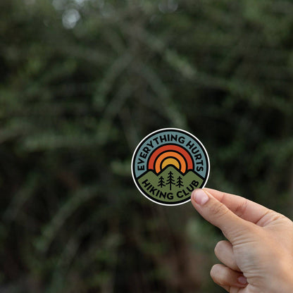 A hand pinches the Everything Hurts Hiking Club sticker in front of blurred dark evergreen branches.