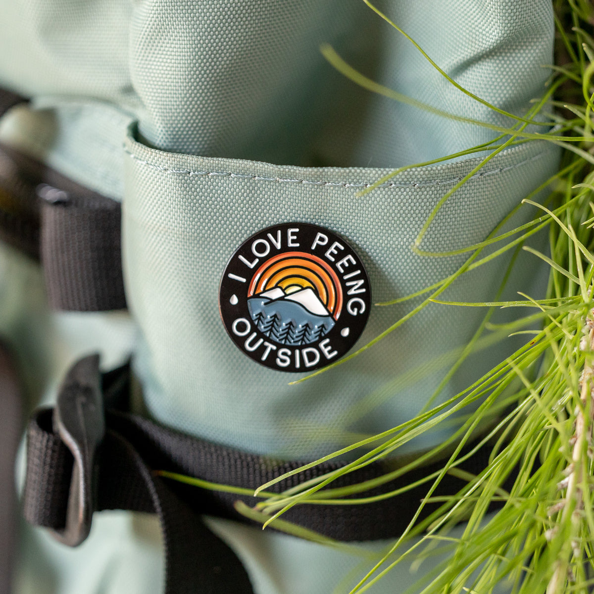 Clipped to a teal pack's front pocket, the I Love Peeing Outside pin sits near tall grass blades and a buckle strap.