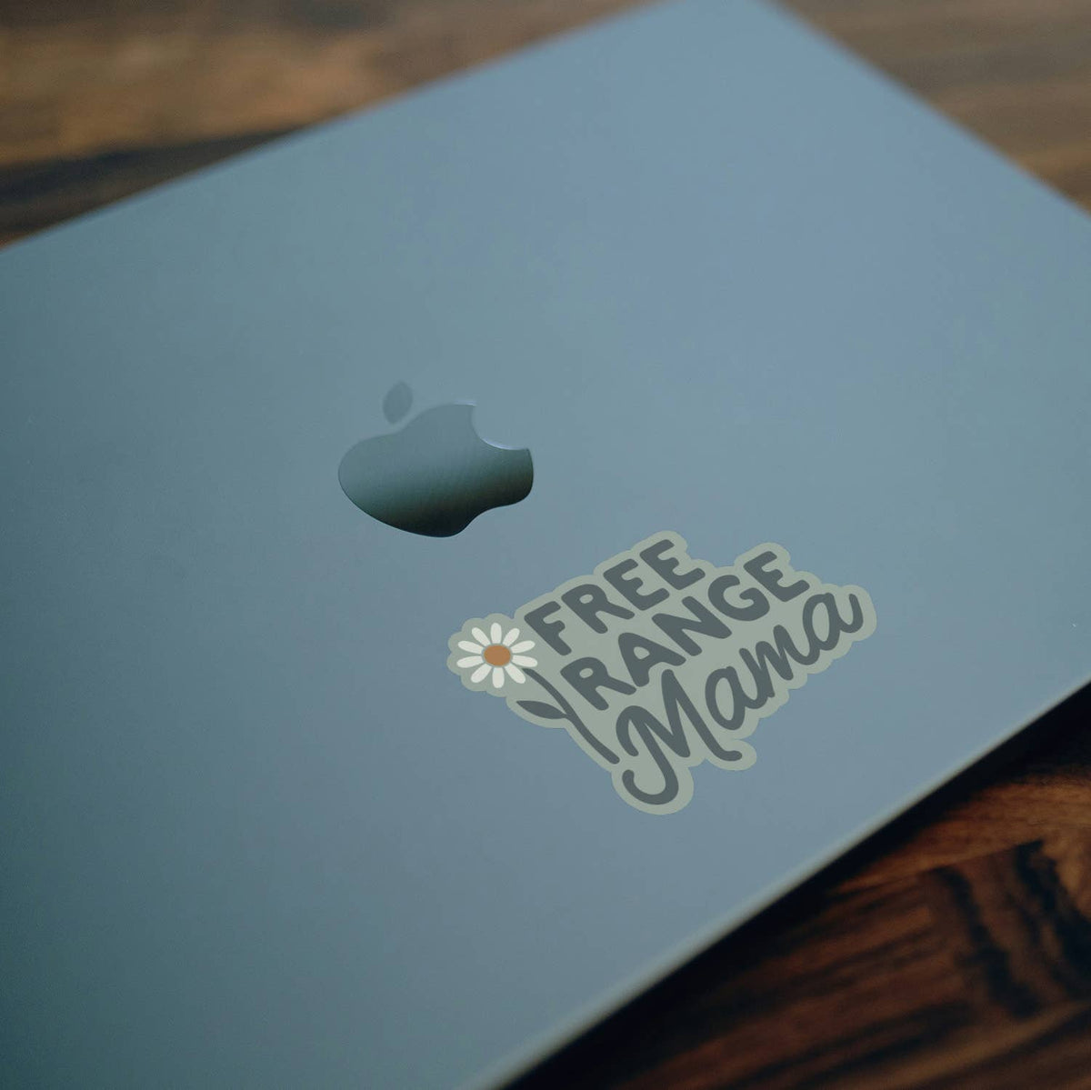 Stuck on the lid of a pale blue laptop, the Free Range Mama sticker sits below the Apple logo on wood.