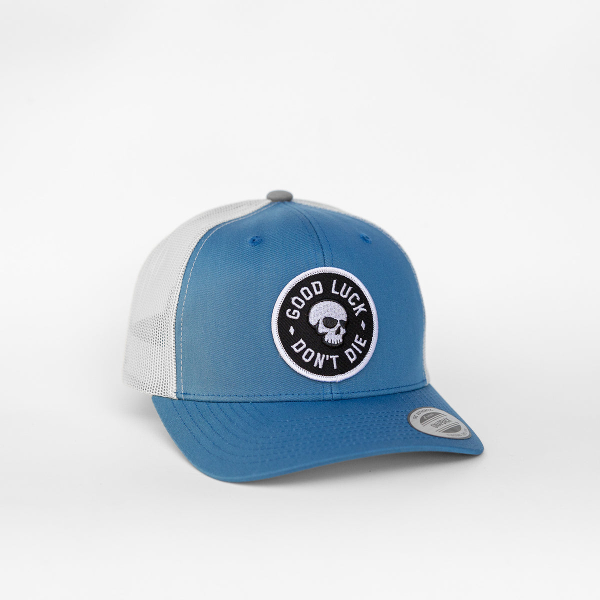 Blue and white panels make up the Good Luck Don't Die trucker hat, its skull patch sitting just above the brim.