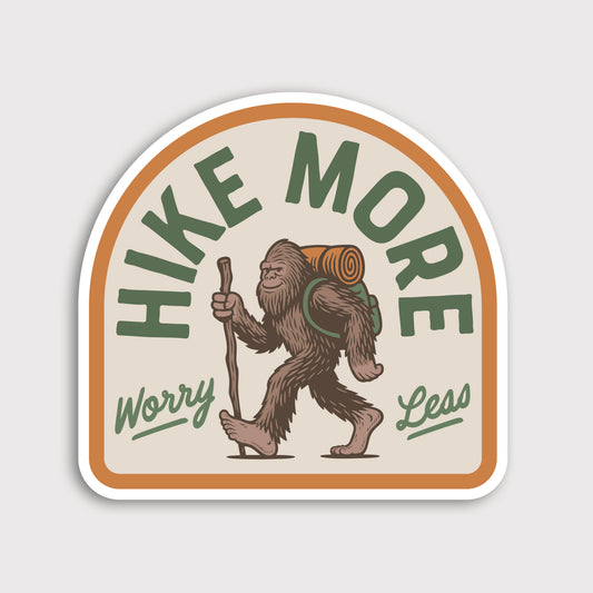 The Hike More Worry Less sticker shows a bigfoot carrying a walking stick and rolled pack on a cream arched background.