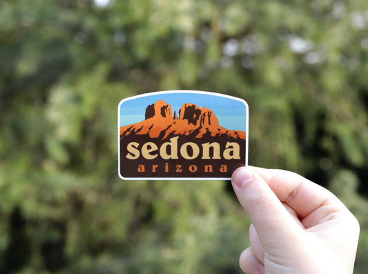 Fingers hold the Sedona Arizona sticker with its orange red rock buttes against blurred green pines.