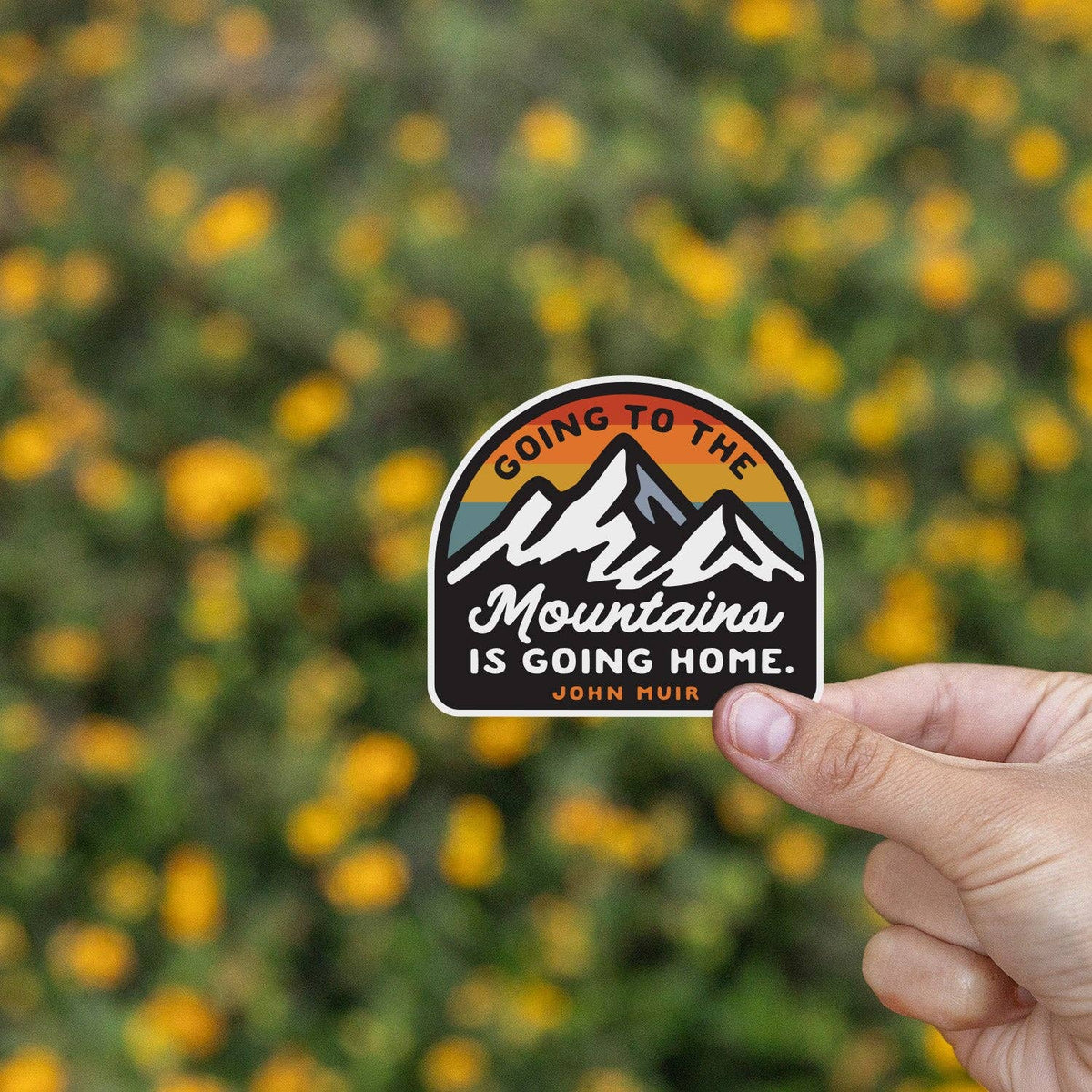 Fingers hold the Going to the Mountains is Going Home sticker before a field of yellow flowers.