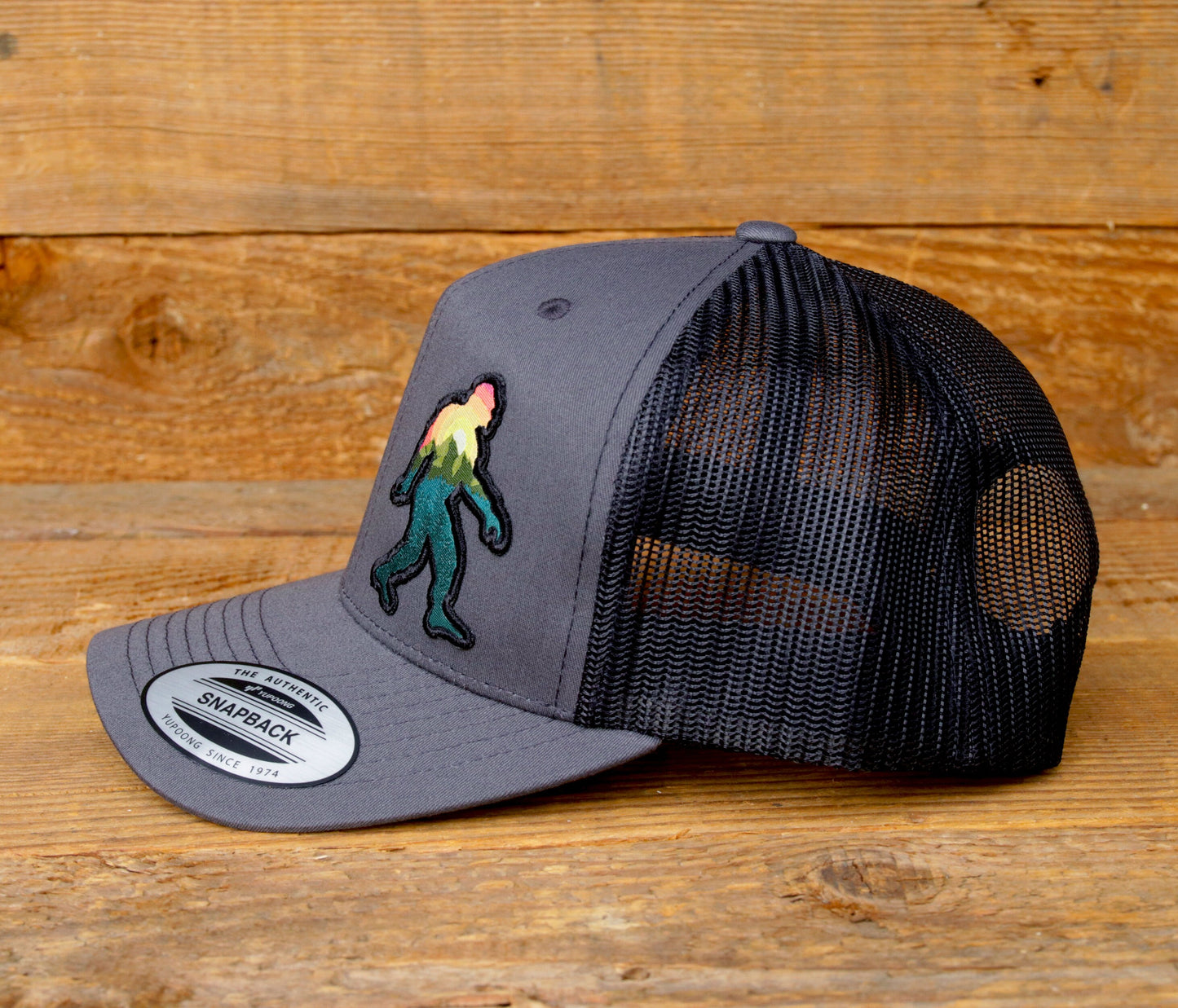 Side profile on barn wood, the grey Bigfoot Adventure trucker hat shows its curved brim and black mesh.