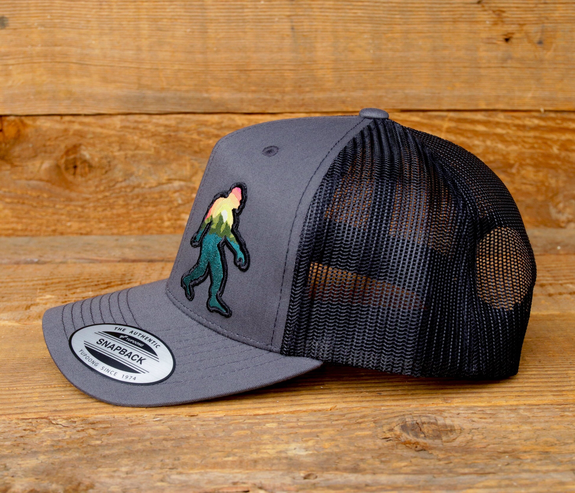 Side profile on barn wood, the grey Bigfoot Adventure trucker hat shows its curved brim and black mesh.