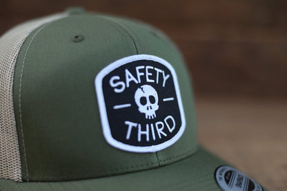 Tight view of the green Safety Third trucker hat where white stitching outlines the skull and lettering.