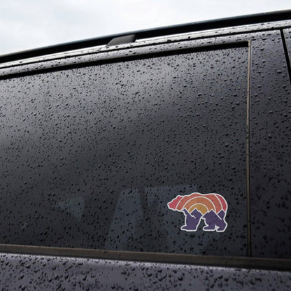 Rain beads across a dark car window where the Adventure Grizzly Bear sticker sits low on the glass.