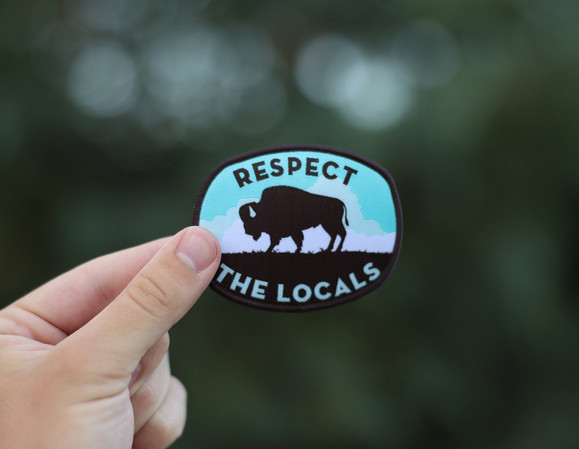 Fingers hold the Respect The Locals bison patch up against soft green trees thrown out of focus.