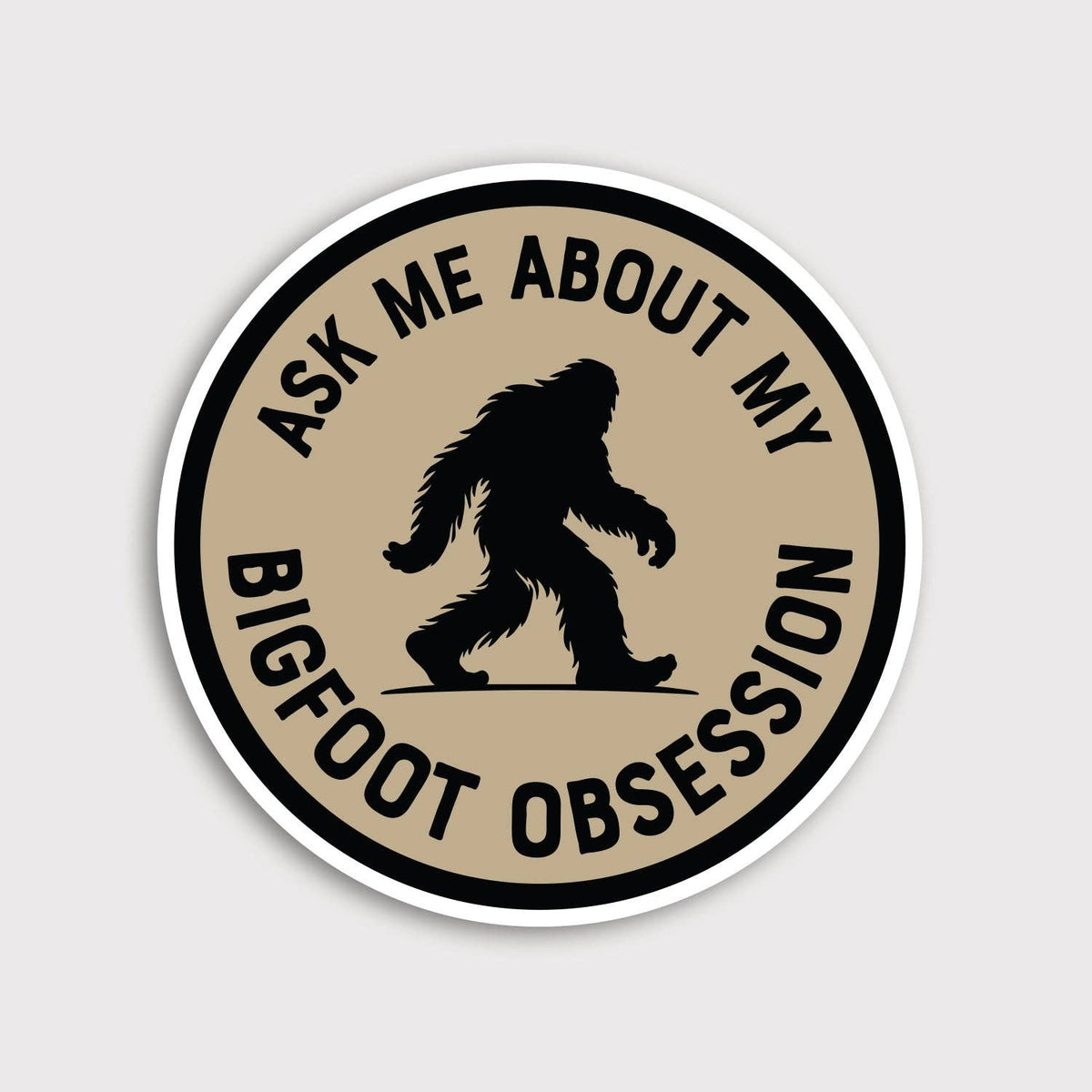Round tan Ask Me About My Bigfoot Obsession sticker with a black Sasquatch silhouette on a plain grey background.