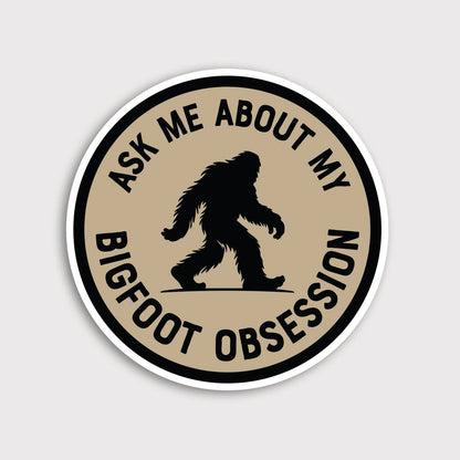 Round tan Ask Me About My Bigfoot Obsession sticker with a black Sasquatch silhouette on a plain grey background.