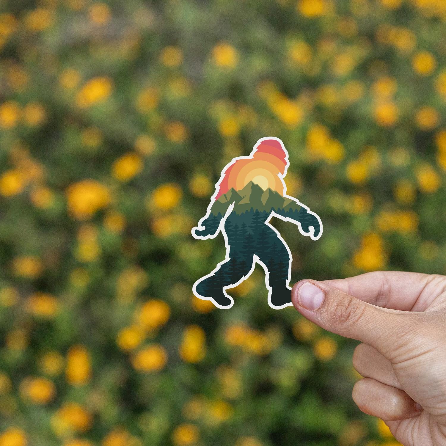 A field of small yellow flowers fills the background as a hand lifts the Bigfoot Adventure sticker.