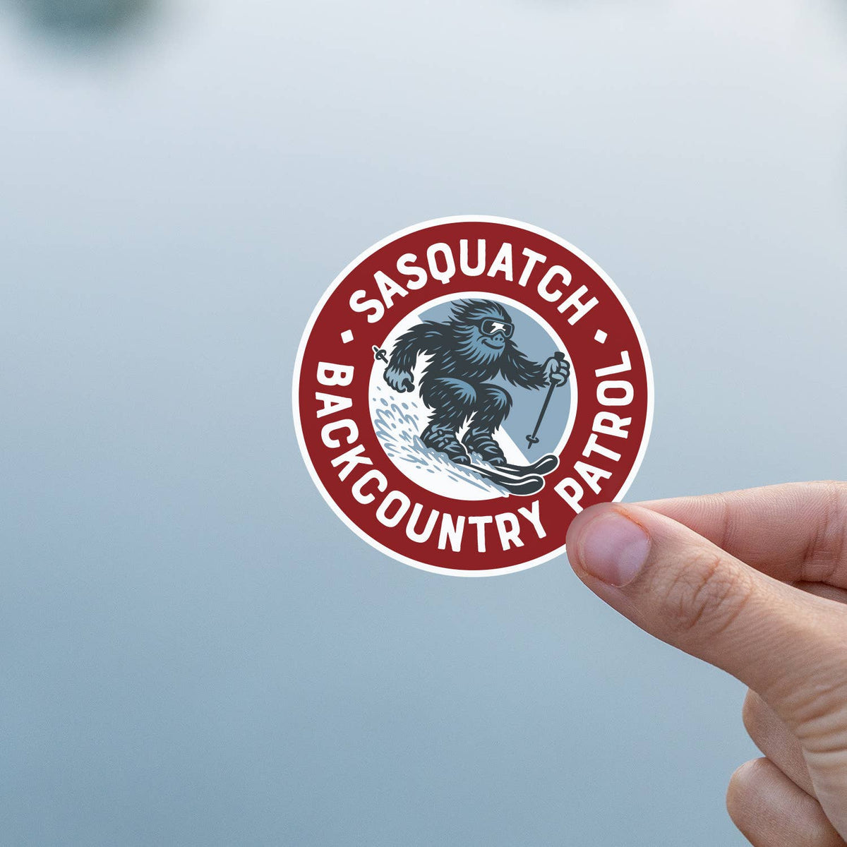 Two fingers hold the Sasquatch Backcountry Patrol sticker against a pale grey blurred background.